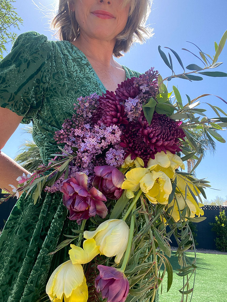 Flower to Table founder outdoors holding a large, vibrant bouquet of flowers including yellow tulips, purple chrysanthemums, lilac, and green olive branches, wearing a textured green dress against a bright blue sky.