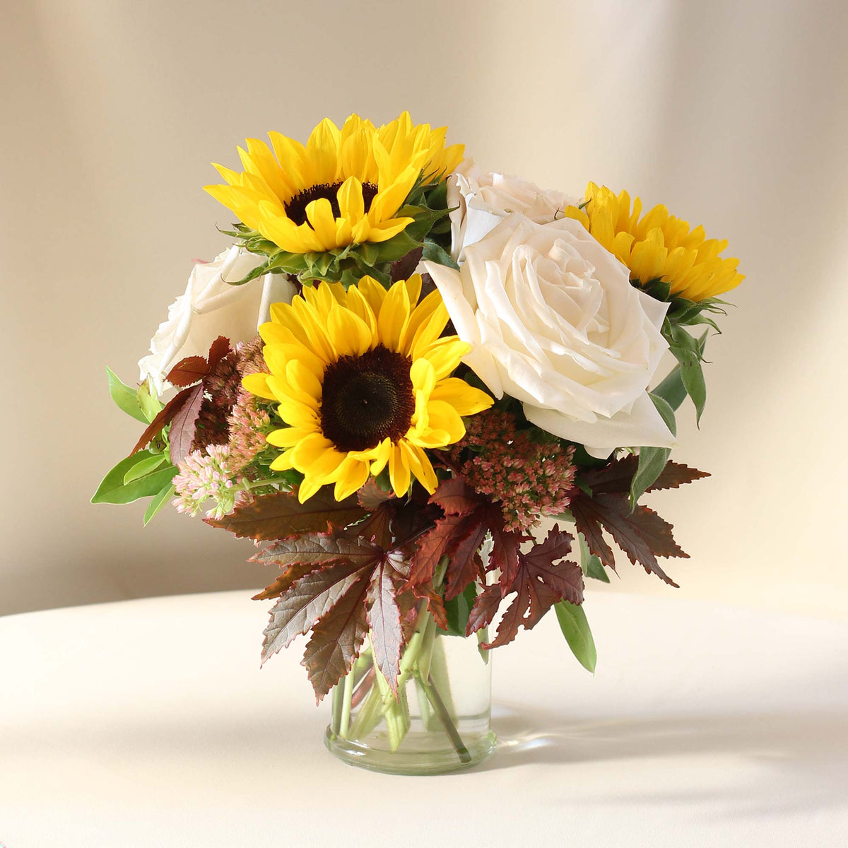 Lil' Ray floral arrangement with yellow sunflowers, white roses, pink sedum and burgundy foliage in a clear glass vase.